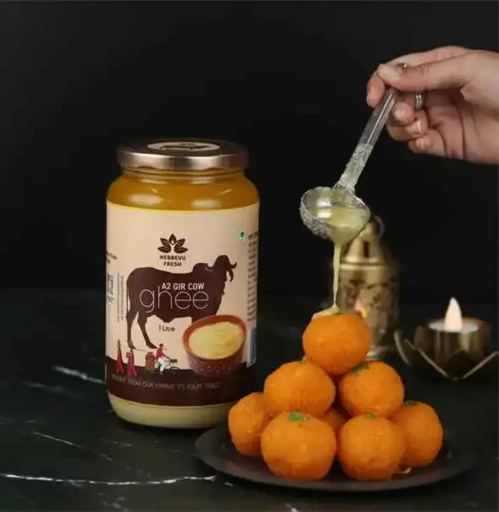 A jar of Hebbevu Fresh A2 Gir cow ghee beside a plate of laddus, with a spoonful of ghee being poured over them by lamplight.