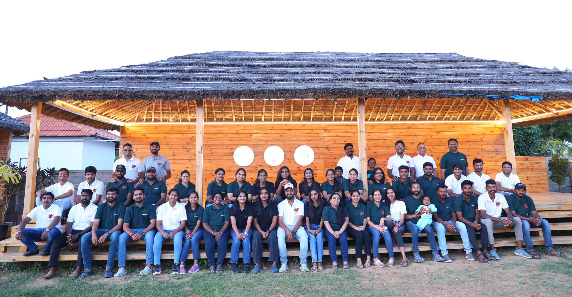 The Hebbevu team, around fifty people in four rows, seated and standing on the steps of the thatched open pavilion at the farm.